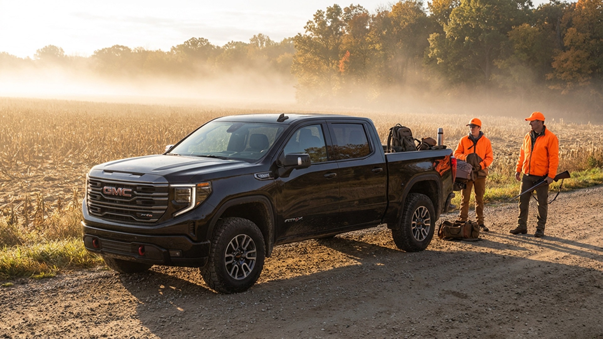Black GMC Sierra AT4X pickup truck parked on a rural dirt road beside a foggy farm field at sunrise while a father and son wearing blaze orange hunting gear prepare equipment during deer season.