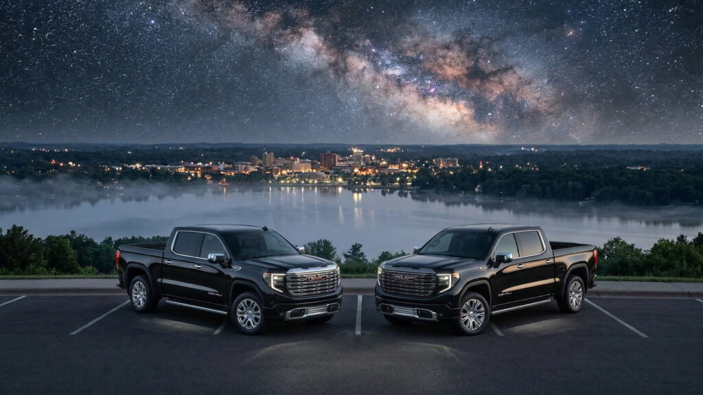 Side-by-side GMC Sierra 1500 Denali and Denali Ultimate trucks parked at night overlooking Lake Wausau with the Milky Way galaxy visible above the city skyline from Rib Mountain in Central Wisconsin.