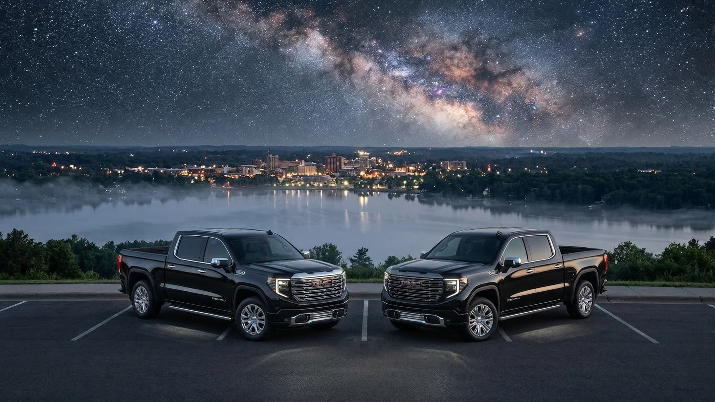 Side-by-side GMC Sierra 1500 Denali and Denali Ultimate trucks parked at night overlooking Lake Wausau with the Milky Way galaxy visible above the city skyline from Rib Mountain in Central Wisconsin.