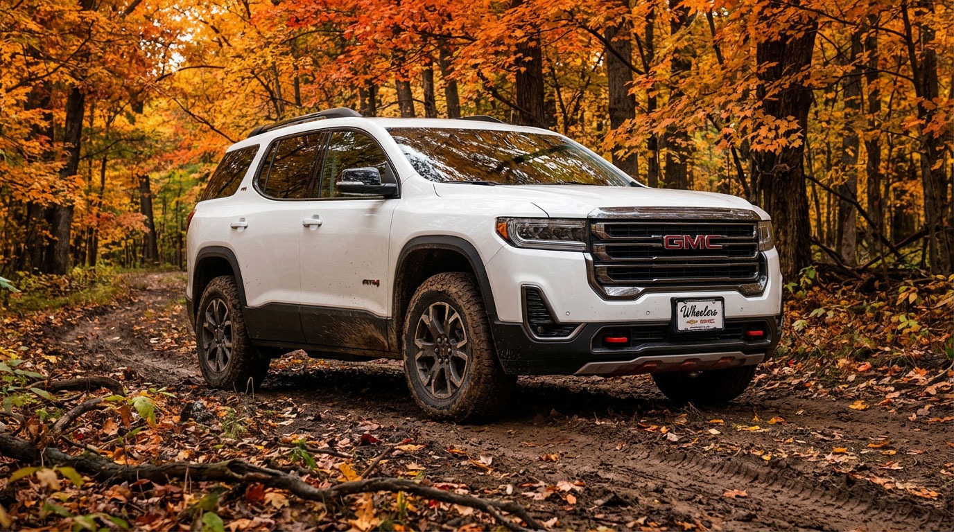 2026 GMC Acadia AT4 on a muddy two-track trail through a Wisconsin hardwood forest in peak fall foliage with orange and red maple canopy overhead and light mud on the lower doors and tires