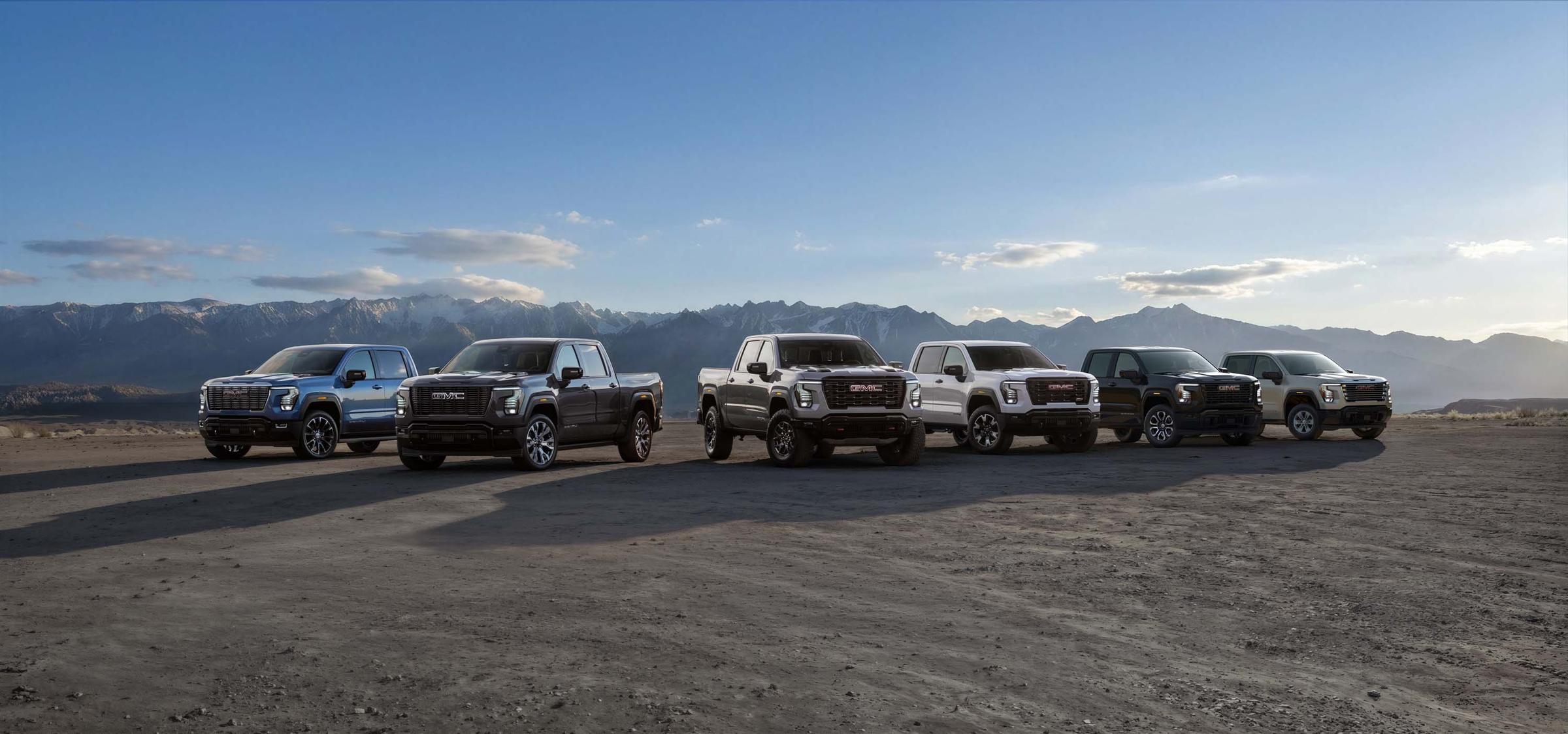 Six 2027 GMC Sierra 1500 pickups parked side by side in front of a mountain range, one of each trim